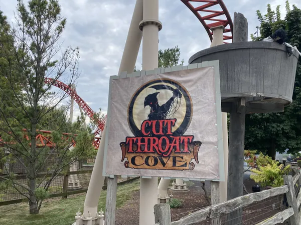 Canvas banner reading Cut Throat Cove hanging from a wooden post at Cedar Point, with coaster track behind it