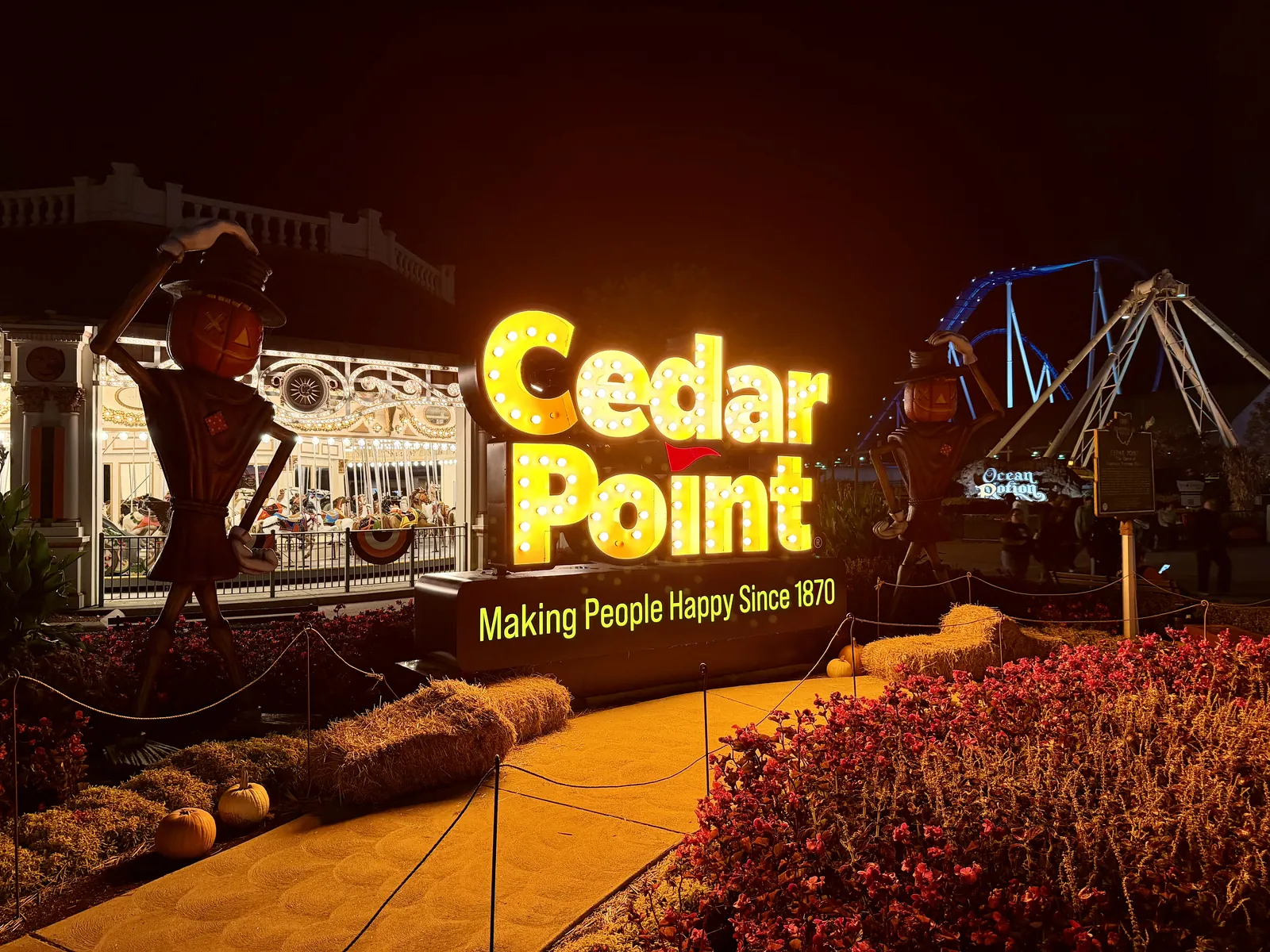 The illuminated Cedar Point marquee at night flanked by pumpkin-headed scarecrows, hay bales and a lit carousel behind it