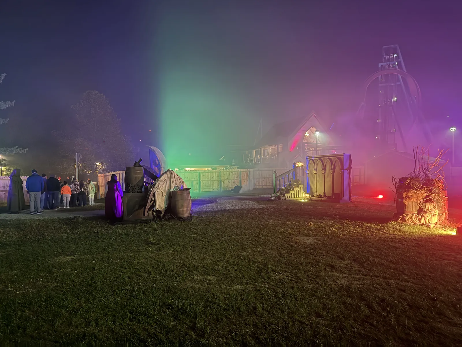 A fog-filled scare zone at night lit green, purple and red, with a robed figure standing beside a queue of guests
