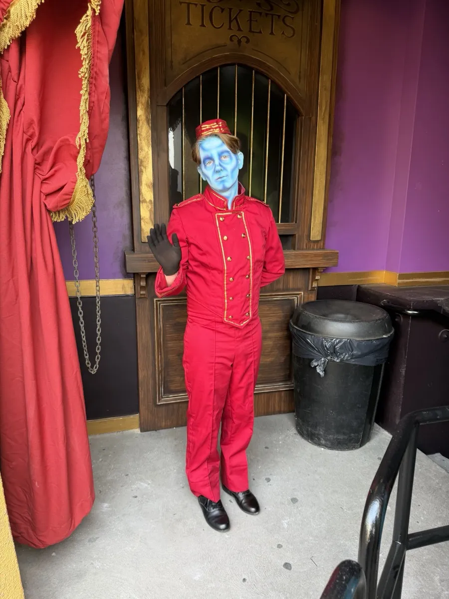 Animatronic bellhop figure in a red jacket standing at a wooden ticket booth in the Phantom Theater queue